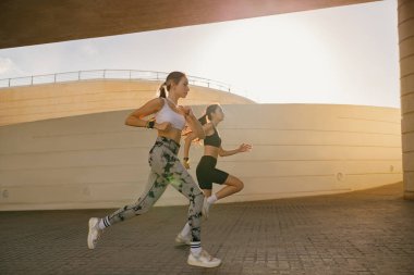 Modern binaların arka planında açık hava pistinde yan yana koşan iki aktif bayan sporcu.