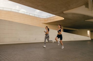 Spor giyimli iki atletik kadın sabahın erken saatlerinde şehirde koşuyor. Hedefe ulaşılıyor.