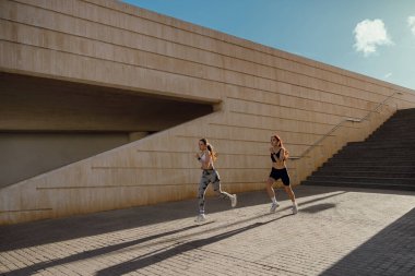 Günesli sabahlarda modern binalarda yan yana koşan iki aktif bayan atlet.