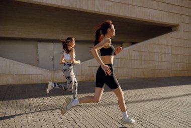 Günesli sabahlarda modern binalarda yan yana koşan iki aktif bayan atlet.