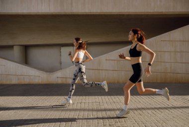 Modern binaların arka planında açık hava pistinde yan yana koşan iki aktif bayan sporcu.