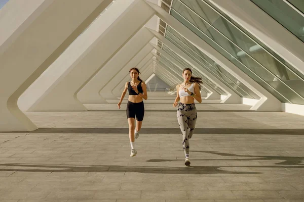 Spor giyimli iki atletik kadın sabahın erken saatlerinde şehirde koşuyor. Hedefe ulaşılıyor.