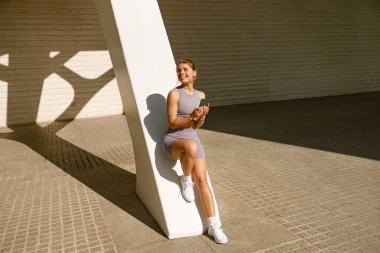 Spor kıyafetleri içinde formda bir kadın eğitimden sonra dışarıda duruyor ve cep telefonu kullanıyor.