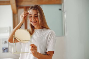 Gülümseyen kadın banyoda aynanın önünde duran pamuklu bezle makyajı temizliyor.