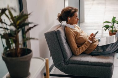 Kadın yönetici, pencerenin yanındaki modern ofiste dizüstü bilgisayarda çalışırken telefonu kullanıyor