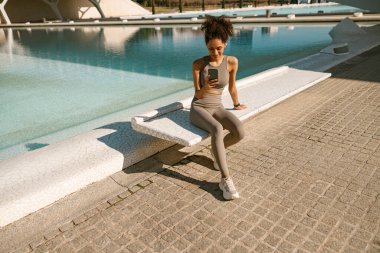 Spor giyim sektöründe genç bir kadın akıllı telefonuyla otururken dışarıda antrenmandan sonra dinleniyor. 