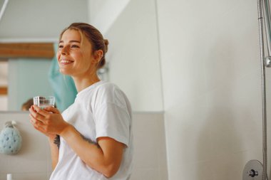 Gülümseyen genç kadın banyoda bir bardak su içiyor. Sabah güzellik rutini