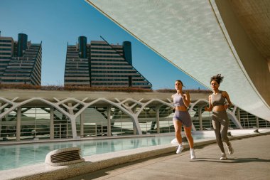 Modern binaların arka planında açık hava pistinde yan yana koşan iki aktif bayan sporcu.