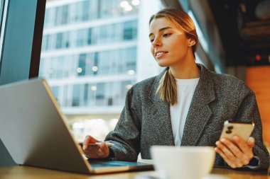 Kadın yönetici telefonda mesaj okuyor ve kafede dizüstü bilgisayarda çalışıyor. Mesafe çalışma kavramı 