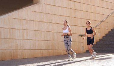 Günesli sabahlarda modern binalarda yan yana koşan iki aktif bayan atlet.