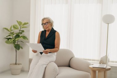 Yaşlı bir kadın sessizce oturuyor ve şık, havadar ve aydınlık bir oturma odasında detaylı bir belge okuyor.