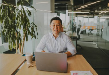 Neşeli, gülümseyen profesyonel bir adam özenle modern ve dinamik bir ofis içinde dizüstü bilgisayar üzerinde çalışıyor.