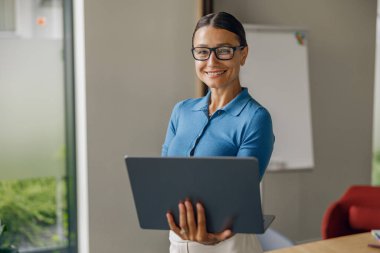 Profesyonel bir kadın, modern ve şık bir ofis ortamında dizüstü bilgisayar kullanırken gülümsüyor.