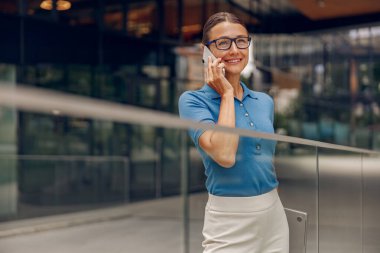 Şık gözlükler takan neşeli bir iş kadını modern bir ofis alanında telefon görüşmesi yapıyor.