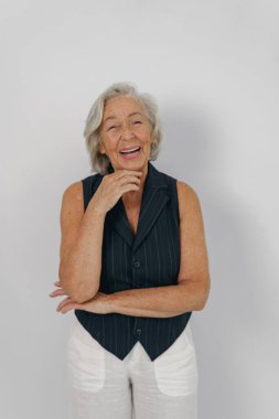 Güzel, gri saçlı yaşlı bir kadın temiz, basit bir arkaplana karşı şık bir kıyafet giyerken sıcak bir şekilde gülümsüyor.