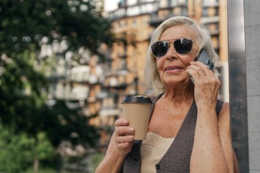 Şık güneş gözlüklü yaşlı bir kadın modern şehir ortamında bir telefon görüşmesi ve sıcak kahveden hoşlanıyor.