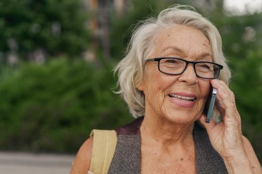 Yaşlı bir kadın, gözlük takarken mutluluk saçıyor. Dışarıdaki yemyeşil alanda canlı bir telefon görüşmesi yapıyor.