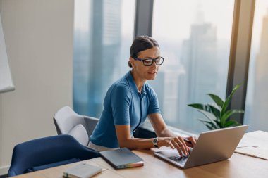 Gözlük takan odaklanmış bir kadın laptopunu şık, modern bir ofis ortamında kullanıyor.