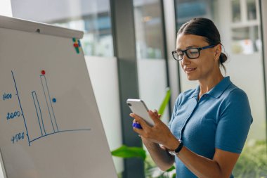 Odaklanmış bir kadın dosyalarla dolu bir beyaz tahtanın önünde dururken akıllı telefonundaki verileri inceliyor.