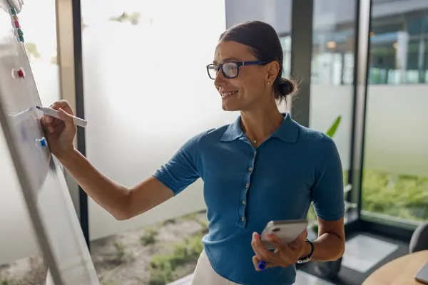 stock image A focused woman in a blue shirt writes on a whiteboard during an important meeting in a modern office