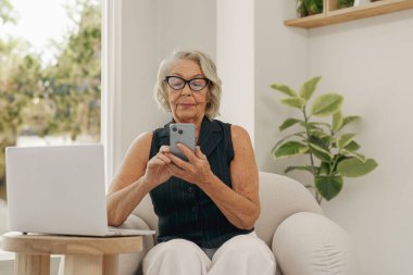 Modern bir ev ortamında, yaşlı bir kadın akıllı telefonuyla rahatça meşgul oluyor, günlük rutininin tadını çıkarıyor.