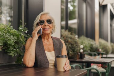 Neşeli, son sınıf öğrencisi, şık güneş gözlüğü takan bir kadın dışarıda telefonla konuşurken kahvesinin tadını çıkarıyor.