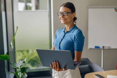 Modern ve şık bir ofis ortamında, profesyonel bir kadın elinde dizüstü bilgisayarla gülümsüyor.