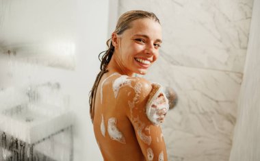 Gülümseyen bir kadın ferahlatıcı bir duşun keyfini çıkarıyor, tamamen sabun köpüğüyle kaplı, saf mutluluk yayıyor.