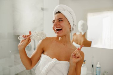 Neşeli bir kadın, havluyla sarılmış, güneşli bir banyoda ferahlatıcı sabah rutininin tadını çıkarıyor.