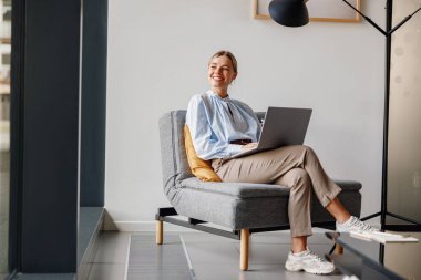 Canlı, genç bir kadın, şık, çağdaş ve davetkar bir ortamda, dizüstü bilgisayarıyla neşe içinde çalışıyor.