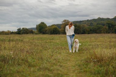 Bir kadın köpeğiyle sakin bir tarlada rahatça dolaşır, doğanın harikalarıyla güzel bir şekilde çevrelenmiş.