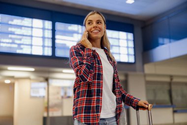 Havaalanında genç bir kadın telefonuyla konuşurken mutlu bir şekilde gülümsüyor. Heyecanlı ve yolculuğuna hazır hissediyor.