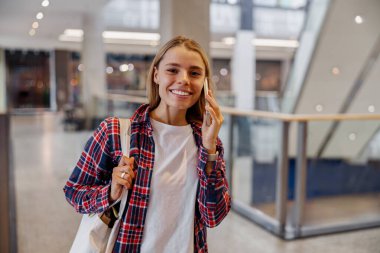 Ekose gömlek giyen genç bir kadın canlı ve çağdaş bir alışveriş ortamında mutlu bir şekilde gülümsüyor.