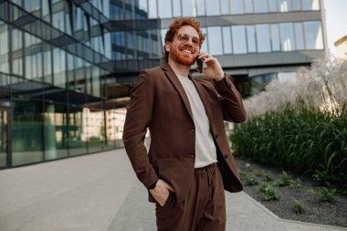 Özel dikim kahverengi takım elbiseli şık bir işadamı etkileyici modern mimarinin dışında bir telefon görüşmesi yapıyor.
