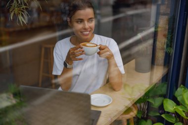 Genç bir kadın laptopuyla çalışırken kafeteryada kahvesinin tadını çıkarıyor, sıradan bir çalışma alanını yansıtıyor..