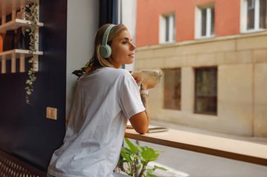 Huzurlu bir sahnede, bir kadın neşeyle müzik dinlerken pencerenin kenarında keyifle kahveyi yudumluyor.