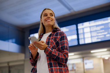 Neşeli genç bir kadın havaalanında akıllı telefonunu tutarak seyahat planlarına hevesle hazırlanıyor.