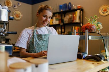 Gülümseyen bir barista enerjik bir şekilde dizüstü bilgisayar kullanırken hareketli bir kahve dükkanı ortamının keyfini çıkarıyor.