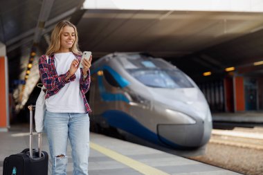 Genç bir kadın, arka planında modern bir tren olan akıllı telefonunu kullanarak tren istasyonunda vakit geçirmekten zevk alıyor..