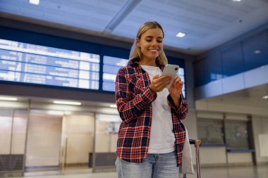 Genç bir kadın, yoğun bir havaalanında akıllı telefonunu kullanıyor. Modern seyahat teknolojisinin faydalarından faydalanıyor.
