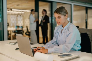 Genç bir kadın dikkatle dizüstü bilgisayarına yoğunlaşarak hareketli ve dinamik bir ofis ortamı yaratıyor.