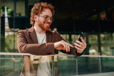 Kıvırcık saçlı neşeli genç bir adam şık ofis ortamında akıllı telefonuyla mutlu bir şekilde iletişim kuruyor.