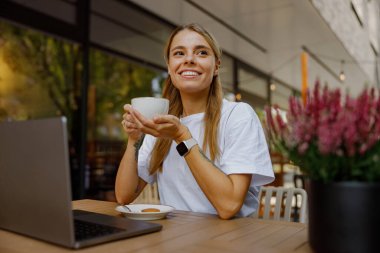 Neşeli bir kadın laptopuyla yaratıcı bir şekilde çalışırken dışarıda kahvesini yudumlamayı sever.