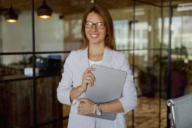 Çevresindeki modern ofis ortamında kendine güvenen bir kadın dizüstü bilgisayar tutuyor.