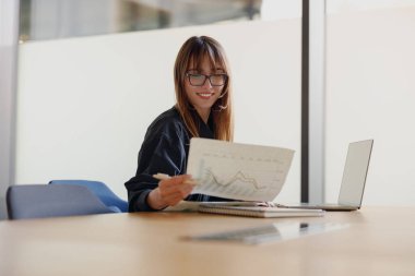 Modern bir ofis ortamında, odaklanmış bir kadın bilgisayarını kullanırken çeşitli veri tablolarını özenle gözden geçirir.