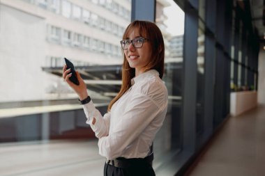 Şık iş kıyafetleri içinde şık bir kadın şık, modern bir ofiste akıllı telefonuyla mutlu mesut meşgul.