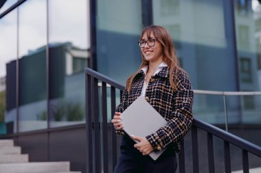 Gözlüklü şık bir genç kadın dışarıda geziyor, elinde parlak bir laptop, modern şehir yaşamını somutlaştırıyor.