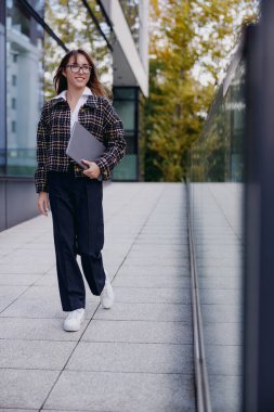 Şık bir kadın canlı bir şehirde dizüstü bilgisayarla, modern profesyonelliği sembolize ederek rahatça yürüyor.