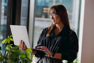 Son derece odaklanmış bir kadın dikkatle dizüstü bilgisayar kullanıyor. Parlak, modern ofis alanındaki canlı bitkilerle çevrili.