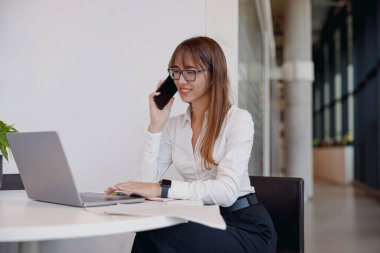 Kararlı bir iş kadını, bilgisayarını parlak ve modern bir ofiste ustalıkla çalışırken telefonla konuşuyor.